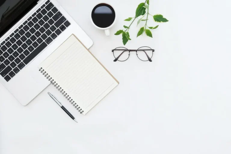 Weisser Schreibtisch von oben fotografiert mit Notebook Notizbuch Kugelschreiber Kaffee Brille und gruener Pflanze